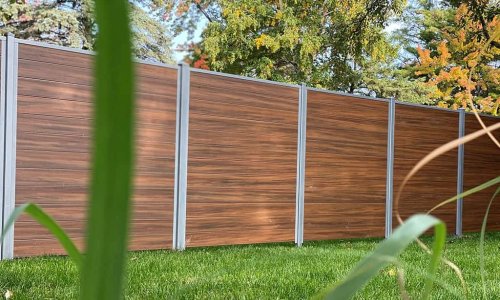 “Wood-grain PVC fence panels with grey posts installed in a residential outdoor space, with grass and trees in the background.”