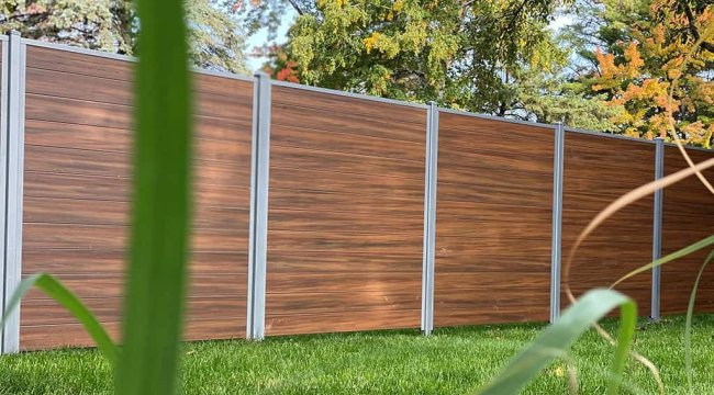 “Wood-grain PVC fence panels with grey posts installed in a residential outdoor space, with grass and trees in the background.”